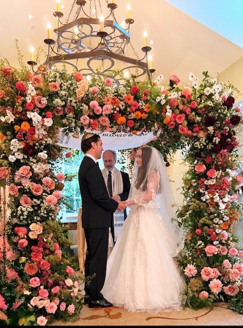 Bride and groom under a floral chuppah with Rabbi Sandy